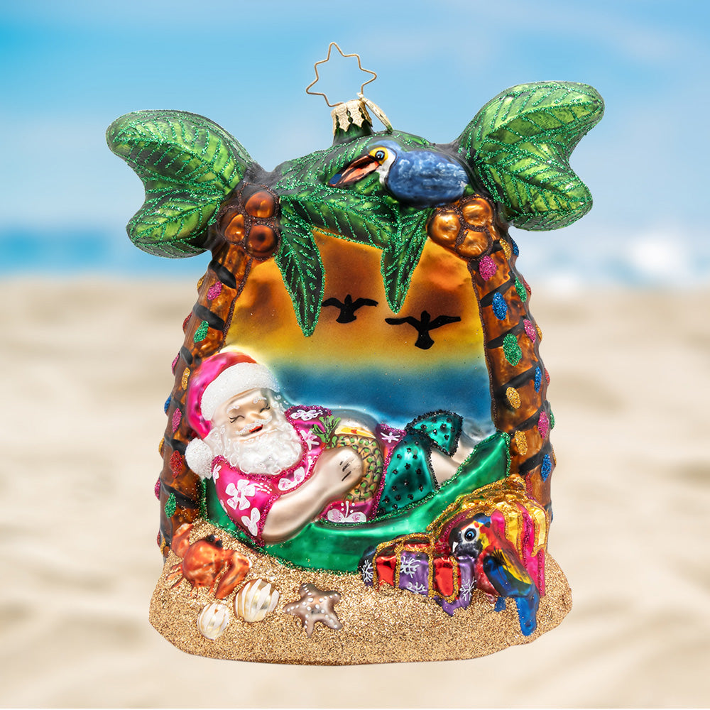 Santa's Breezy Break. Santa in a pink tropical shirt, laying in a green hammock under two decorated palm trees, surrounded by 5 birds and presents and a sunset for a background. View of front