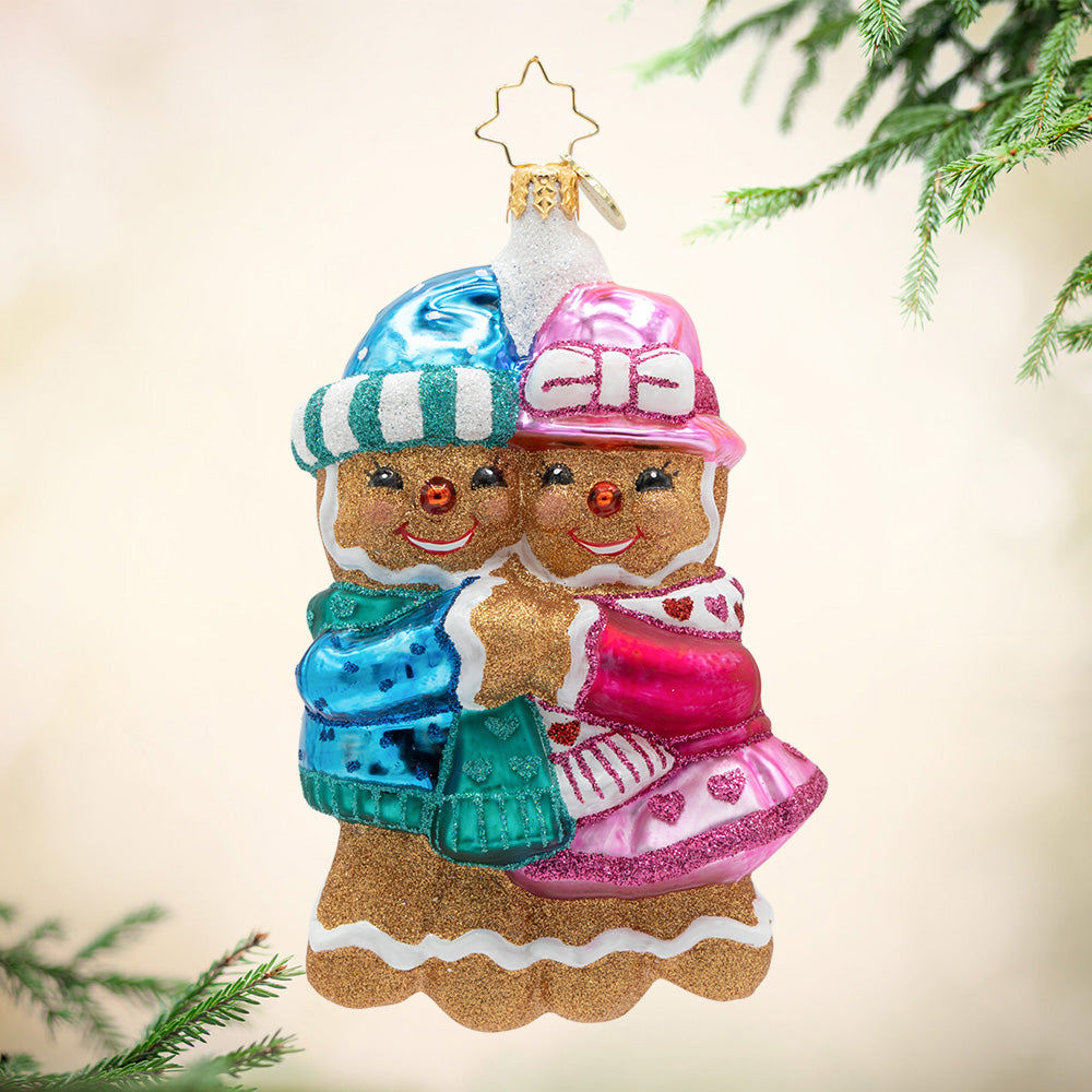 Gingerbread Sweethearts. Ornament of a gingerbread man dressed in a matching blue sweater, scarf and robe hugging a gingerbread girl dressed in a matching pink hat, scarf, and coat. View of front