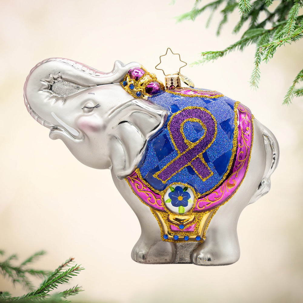 Trunk of Memories. A silver elephant ornament wearing a blue and pink with gold details robe with the Alzheimer's ribbon. View of front