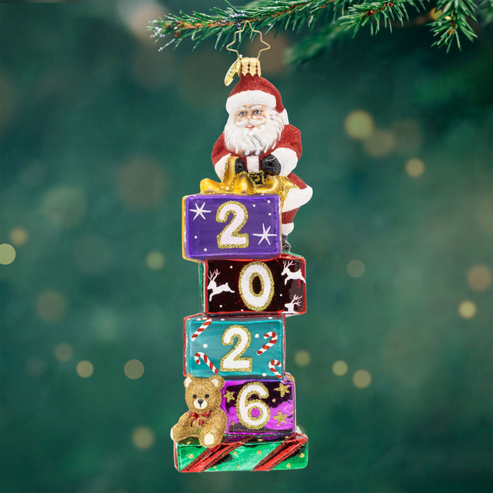 2026 Stacking Up The Years. Ornament of Santa stacking presents in the colors purple, red, blue, and green with the year 2026. View of front