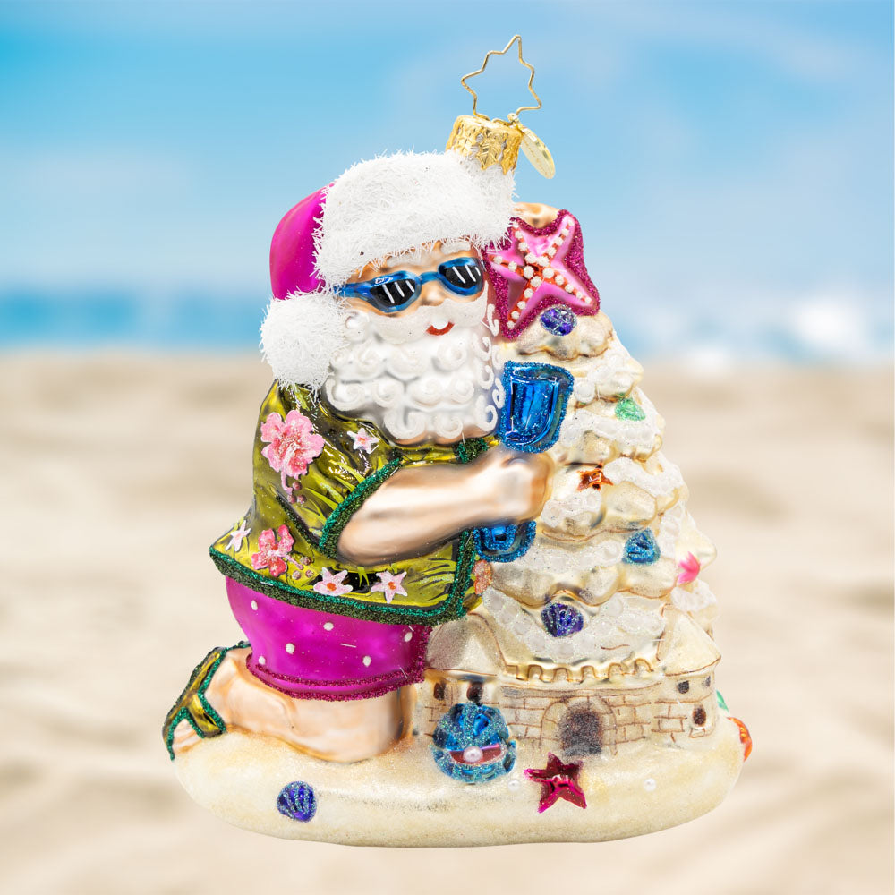 Sandy Claus Castle. Ornament of Santa, in a green tropical top and pink matching shorts and hat, while making a sandcastle with a blue shovel. view of front