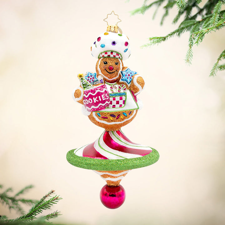 Sweet Winter Whirl. Ornament of a gingerbread man wearing a white hat and apron with gems while holding a jar of cookies with a base in the shape of a spinner in pink, white, and green stripes. View of front