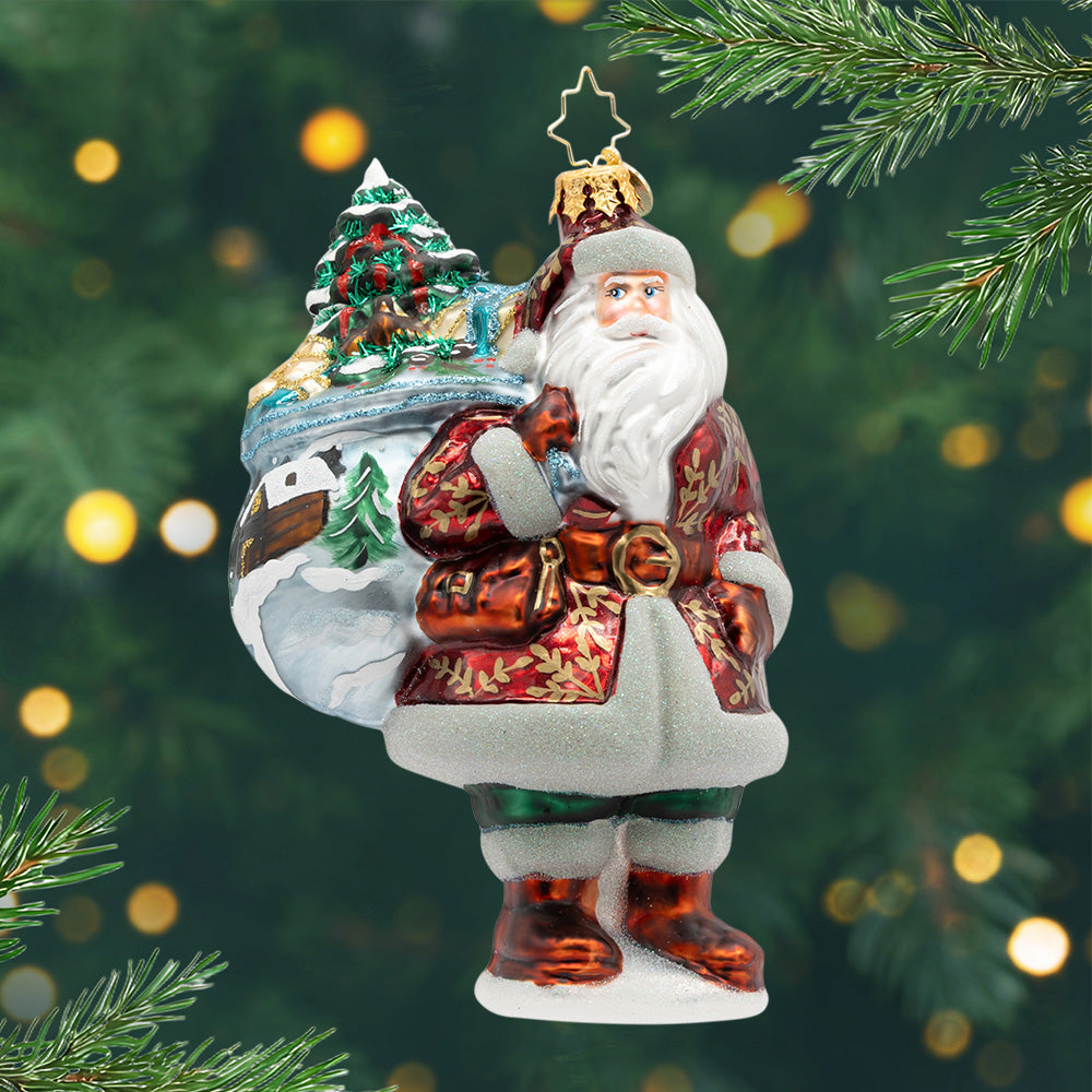 Woodland Noel. Santa ornament of him in a red suit with gold leaf patterns and holding a bag painted with a log cabin scene along with a tree inside the bag. View of front