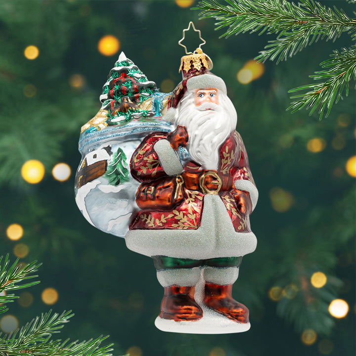 Woodland Noel. Santa ornament of him in a red suit with gold leaf patterns and holding a bag painted with a log cabin scene along with a tree inside the bag. View of front