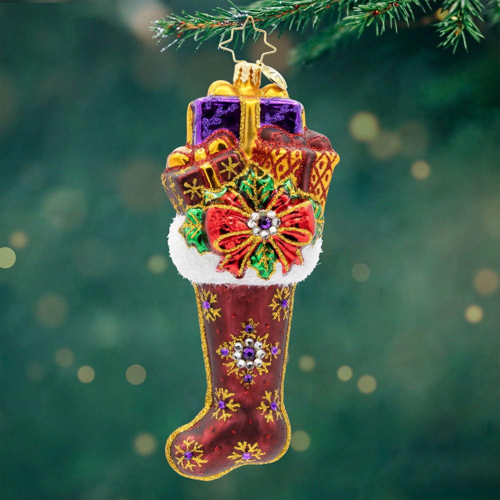 The Gilded Stocking. Red stocking ornament with gold trim and purple gems along with golf and purple presents popping out with a mistletoe and red bow on the top. View of front