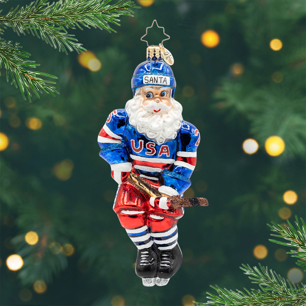 Hat Trick Holiday. Ornament of Santa in a blue and red hocky uniform with the phrase "USA." View of front