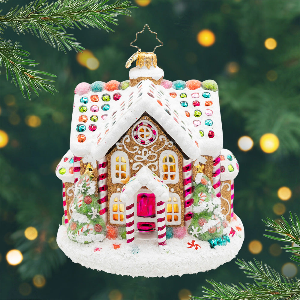 Sweet Treat House.  A gingerbread house ornament with a white snowy roof and colorful gems and five golden windows in the front and to decorated Christmas trees. View of front
