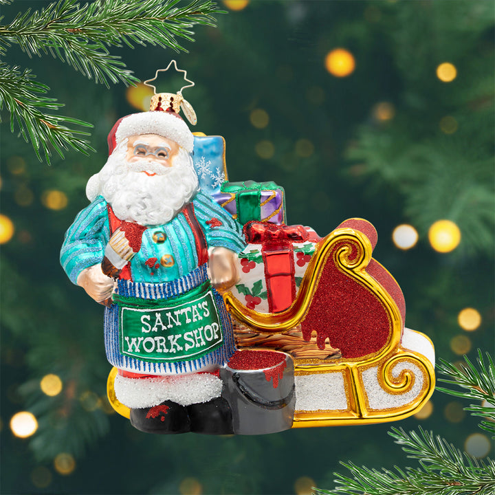 Color Me Christmas! Ornament of Santa in a teal sweater with a blue stripped apron with the phrase "Santa's Workshop" and painting his present filled sleigh red. View of front