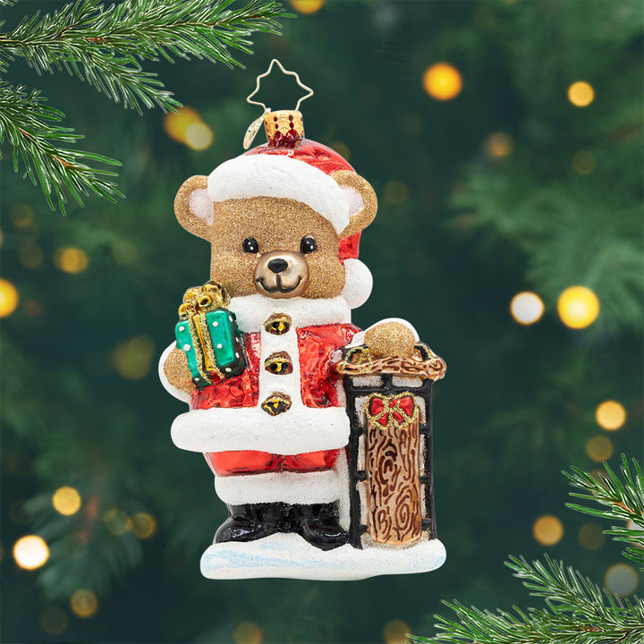 Beary Merry Christmas. Brown bear ornament dressed in a red Santa suit while holding a green present and leaning on a wooden post. View of front