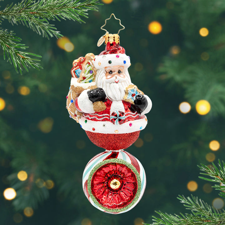 Sweet Claus Reflector. Sparkly ornament of Santa holding a brown bag of presents on top of another ornament with a stripped pattern of red, white and blue with a red center. View of front.