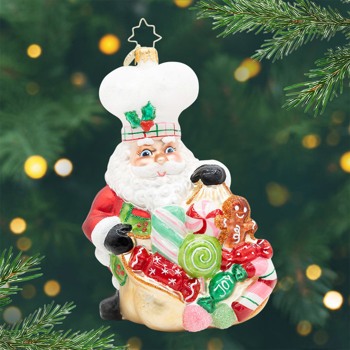 Santa's Sweetest Batch. Ornament of Santa wearing a white chef hat and holding a bag full of green, red, and pink candy. View of front