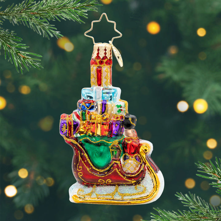 Sleighful of Surprises Gem. A red with gold detailed sleigh ornament carrying a green bag full of stacked, colorful presents with a nutcracker in the front. View of front