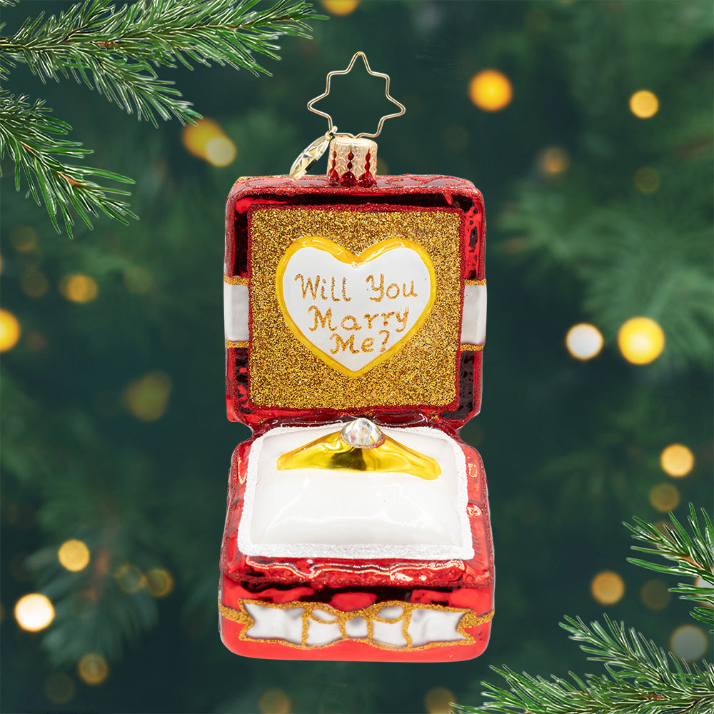 Loves Sparkling Promise Gem. A red, gold, and white ring box ornament with a golden ring in the center and the phrase "Will You Marry Me?" View of front 
