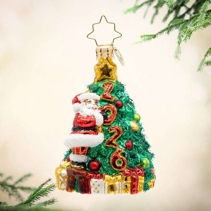 Santa's 2026 Tree Trim Gem. Ornament of Santa next to a decorated Christmas tree with the year 2026 in red and gold and presents surrounding the bottom. View of front