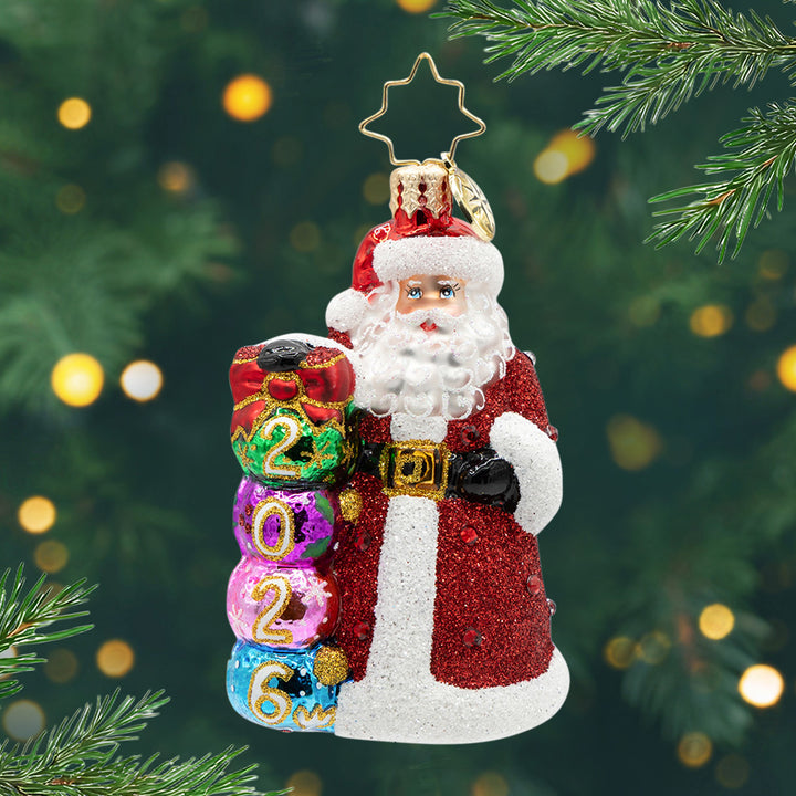 2026 Christmas Countdown Gem. Santa dons a glittering red jeweled coat.  Ornaments with "2026" written on them are stacked next to his side. View of front.