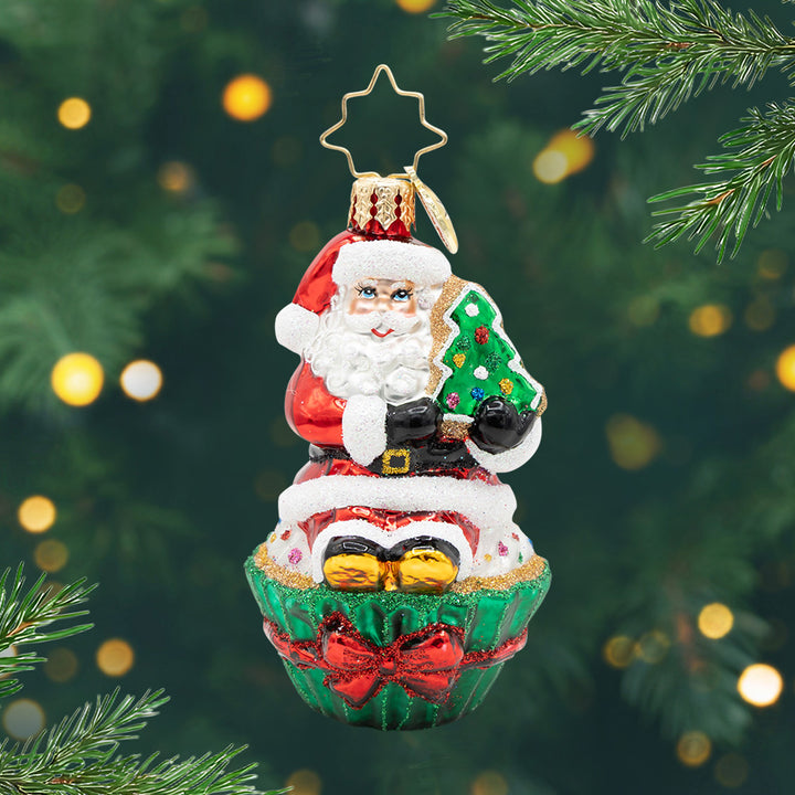Cupcake Claus Gem. Ornament of Santa in his classic red suit sitting on a cupcake with a green wrap and a red bow and holding a Christmas tree. View of front.