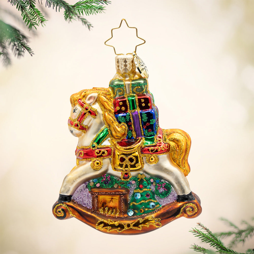 Rockin' Around Christmas Gem. Sparkling ornament of a white rocking horse with a golden mane holding colorful presents of red, blue and green with a brown fireplace and a decorated Christmas tree beneath it. View of front