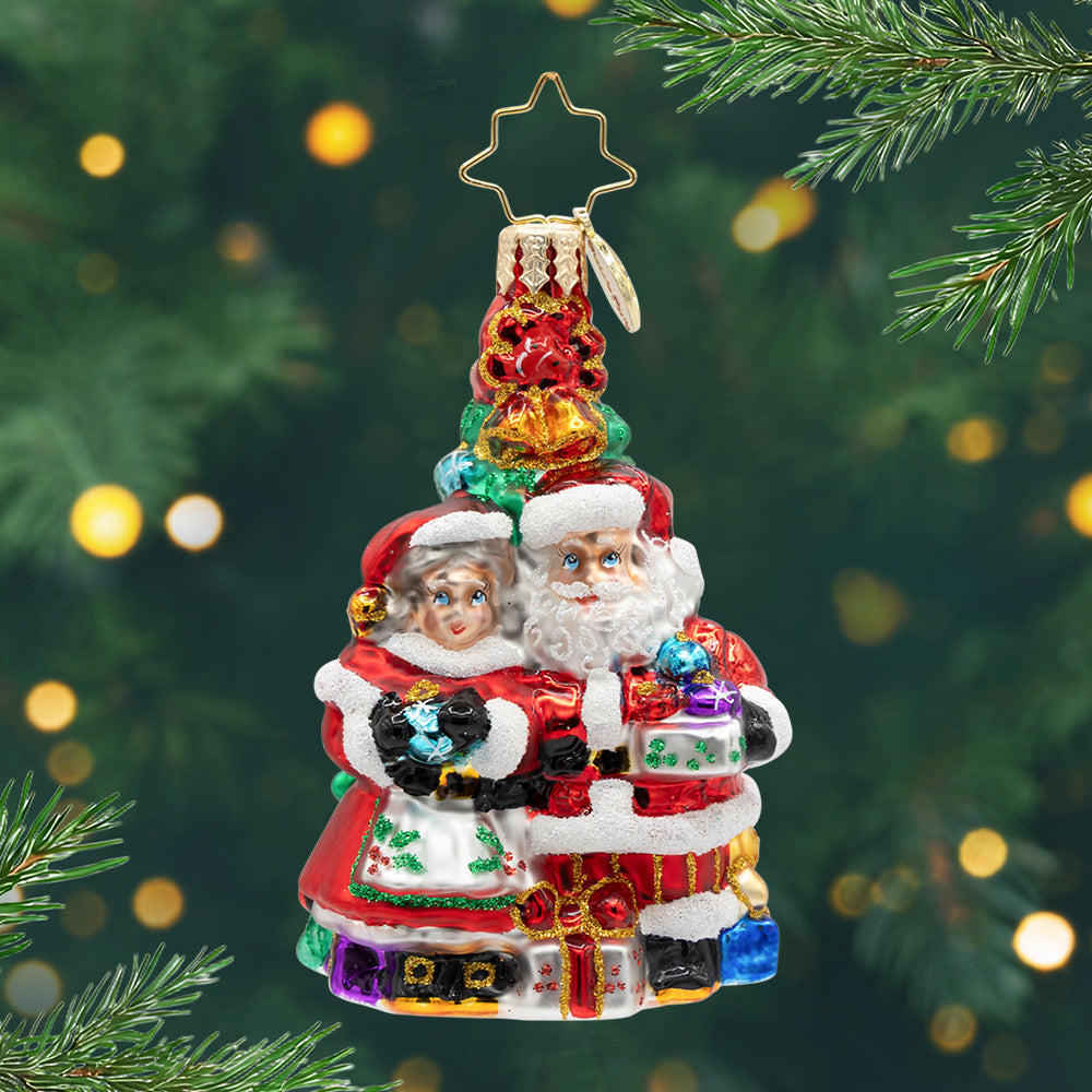 Holiday Harmony Gem. Mr. and Ms. Clause ornament of them in their classic red outfits standing in front of a Christmas tree and holding presents. View of front