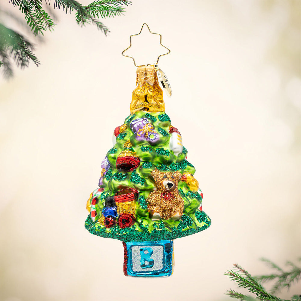 Bundle of Joy Spruce Gem. A Christmas tree ornament decorated in baby toys featuring a teddy bear, train, bottle, and a blue block with the letter B for a stump. View of front