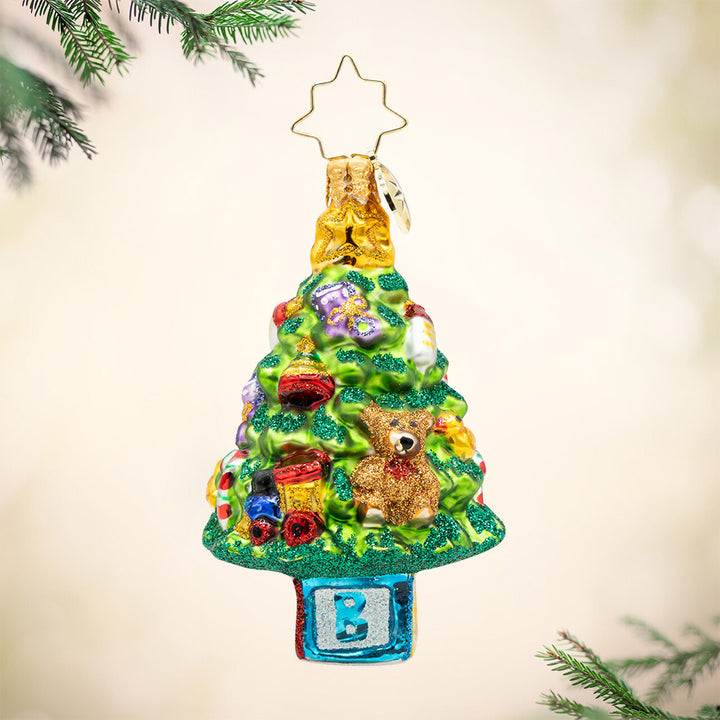 Bundle of Joy Spruce Gem. A Christmas tree ornament decorated in baby toys featuring a teddy bear, train, bottle, and a blue block with the letter B for a stump. View of front
