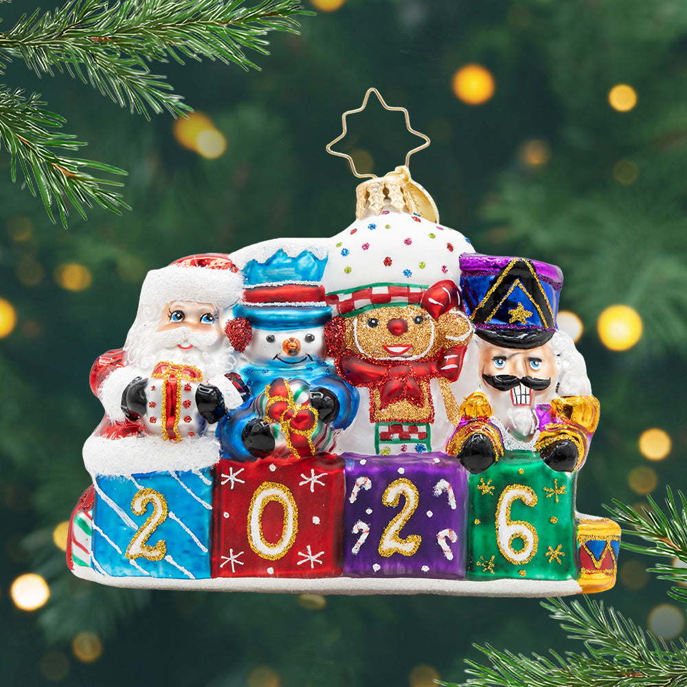 Stacking Up The Cheer Gem. Ornament of Santa, a snowman, gingerbread man, and a nutcrackers, with the colors red, blue, and purple, popping out of four presents that spell out the year 2026. View of front