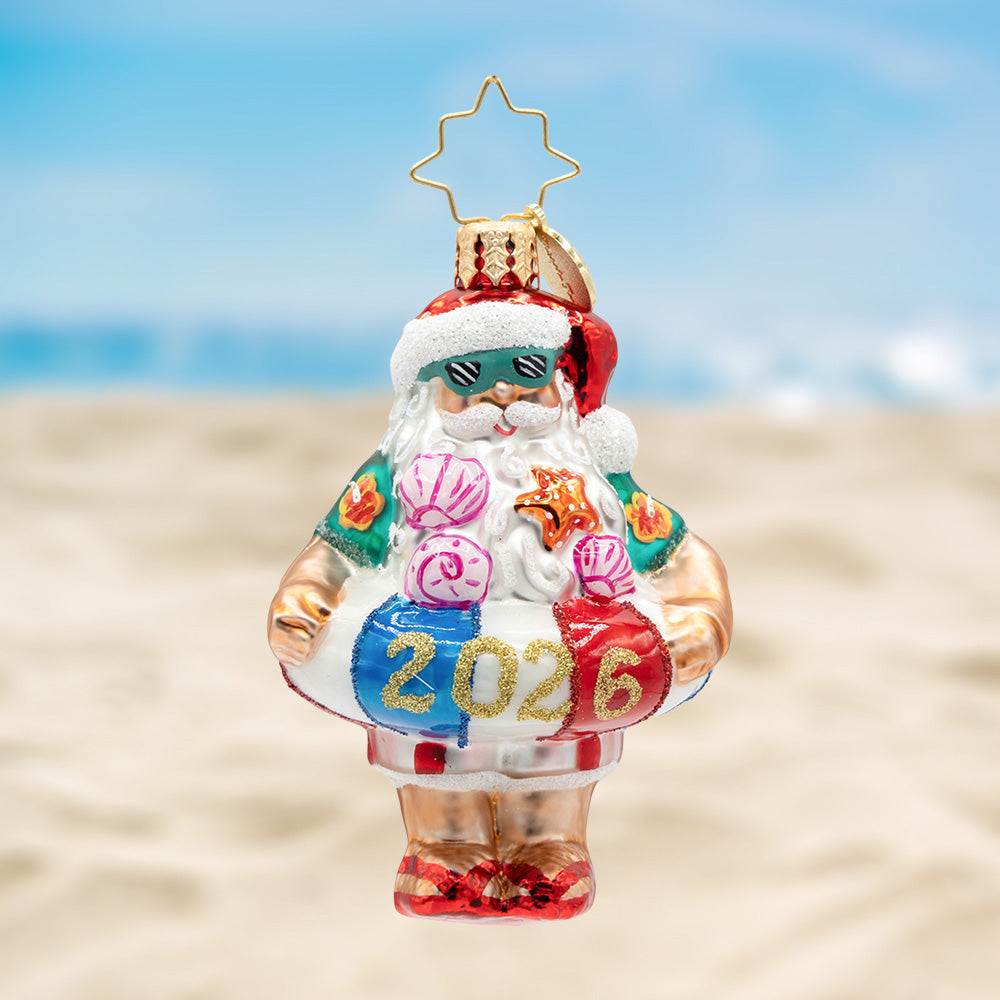 Sun-Kissed Klaus Gem. Ornament of Santa in a tropical green shirt and wearing a red, white, and blue floaty with the year 2026 in gold. View of front
