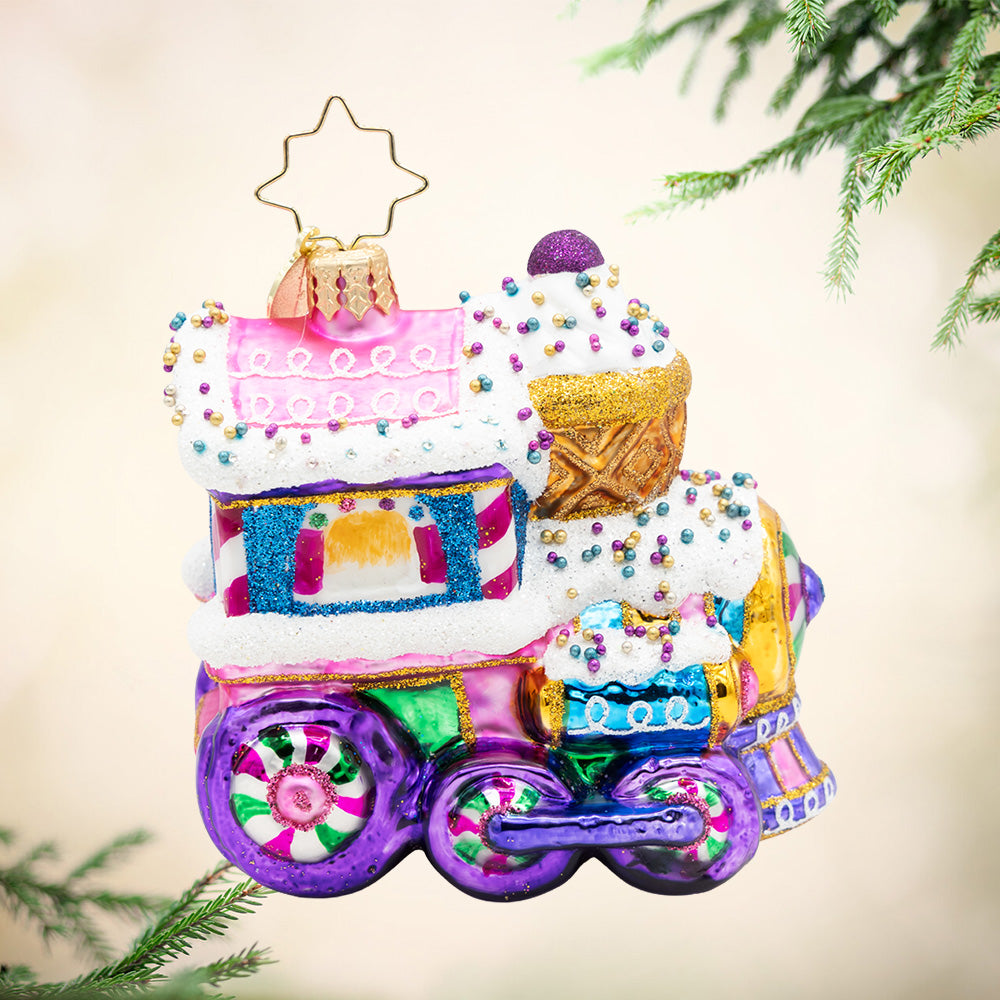 Sugar Rush Express Gem. A train ornament with colors of purple, pink, blue, and gold, while covered in snow with colorful gems and detailing. View of front