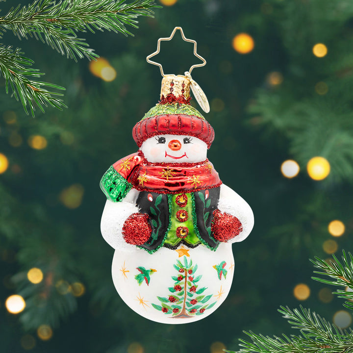 Festive Folk Flair Gem. Ornament of a snowman in a black and green vest with a red and green sparkly hat and matching scarf, along with a tree painted on the bottom. View of front