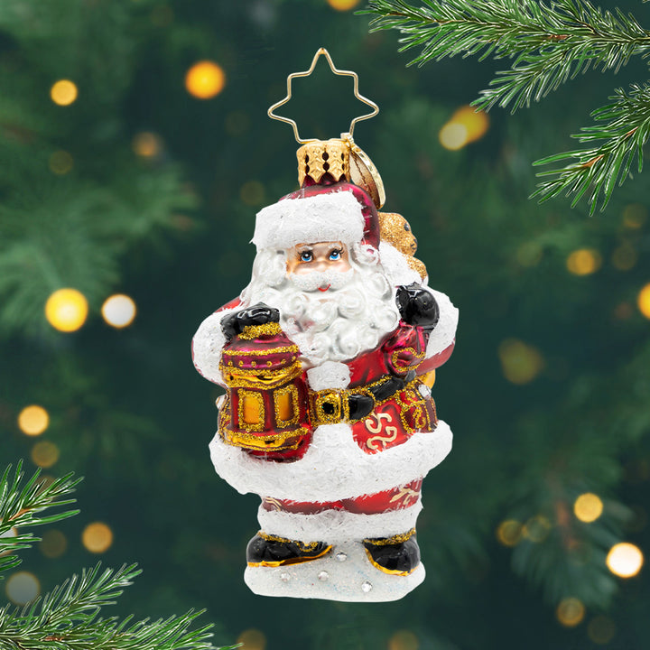 A Claus To Celebrate Gem. Ornament of Santa in red and gold detailed suit holding a bag and a red lantern. View of front