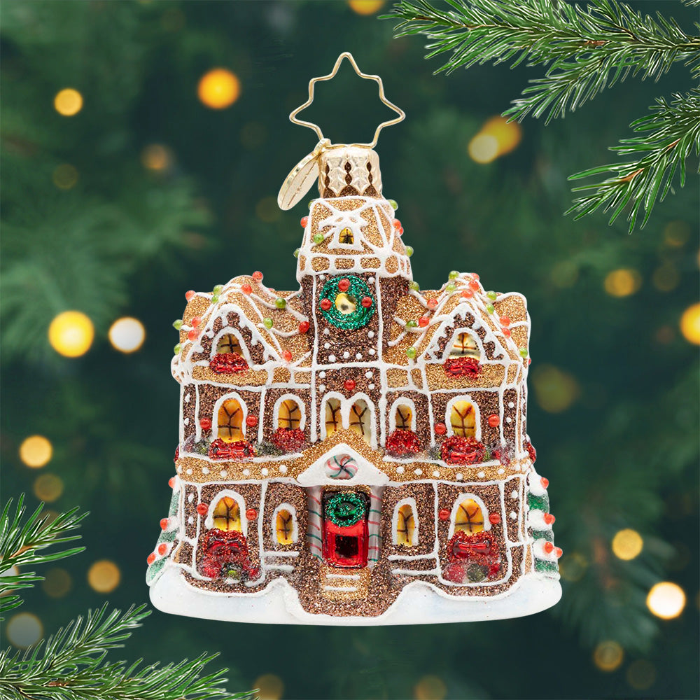 Gingerbread Manor Gem. Ornament of a gingerbread house in a sparkling brown with golden windows and red bows in front of them and a red door. View of front.