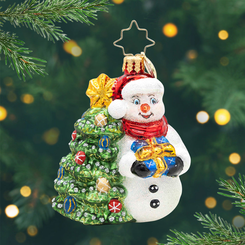Snowy & Bright! Gem. Snowman ornament dressed in a red hat and scarf, holding a blue present next to a decorated Christmas tree. View of front