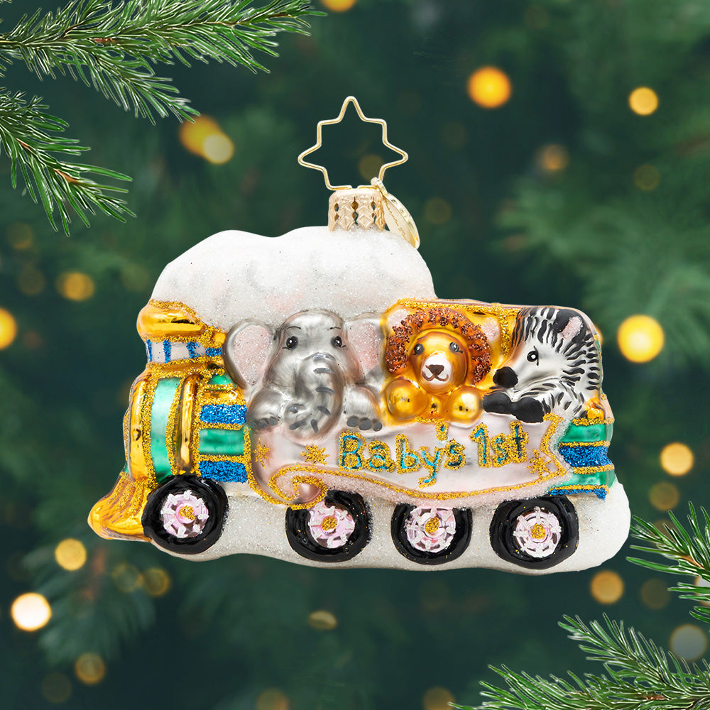 Tiny Travelers Gem. Ornament of teal train with gold details carrying an elephant, a lion, and a zebra with a pink banner with the phrase "Baby's 1st." View of front