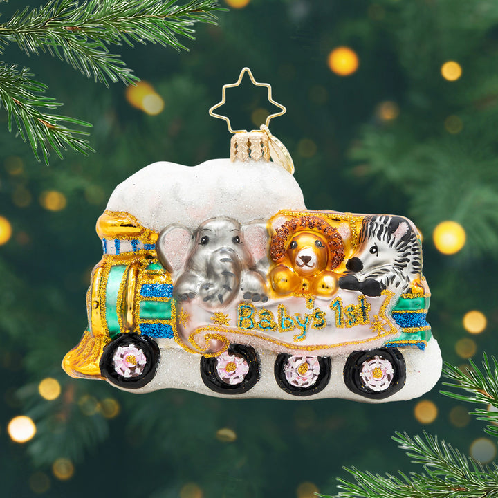 Tiny Travelers Gem. Ornament of teal train with gold details carrying an elephant, a lion, and a zebra with a pink banner with the phrase "Baby's 1st." View of front