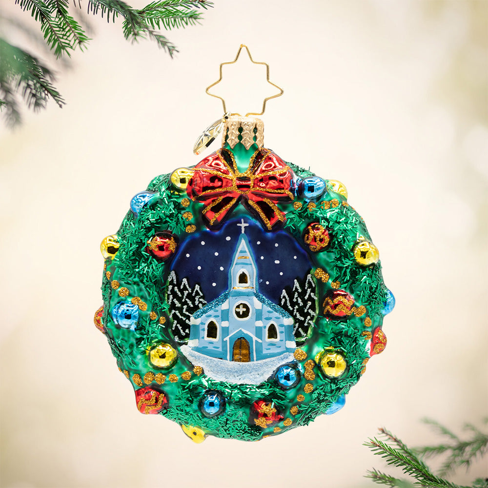 Sacred Harmony Wreath Gem. Wreath ornament decorated in red, yellow, and blue gems, a red bow at the top and a scene of a snow covered church painted in the front. View of front.
