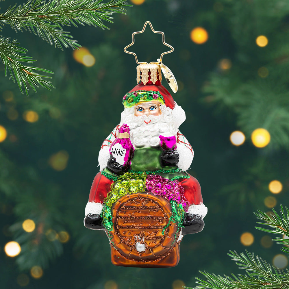 I Believe in Wine Santa Gem. Ornament of Santa in a white sweater with stripes and a green apron sitting on a brown barrel and hold a pink bottle and glass of wine. View of front