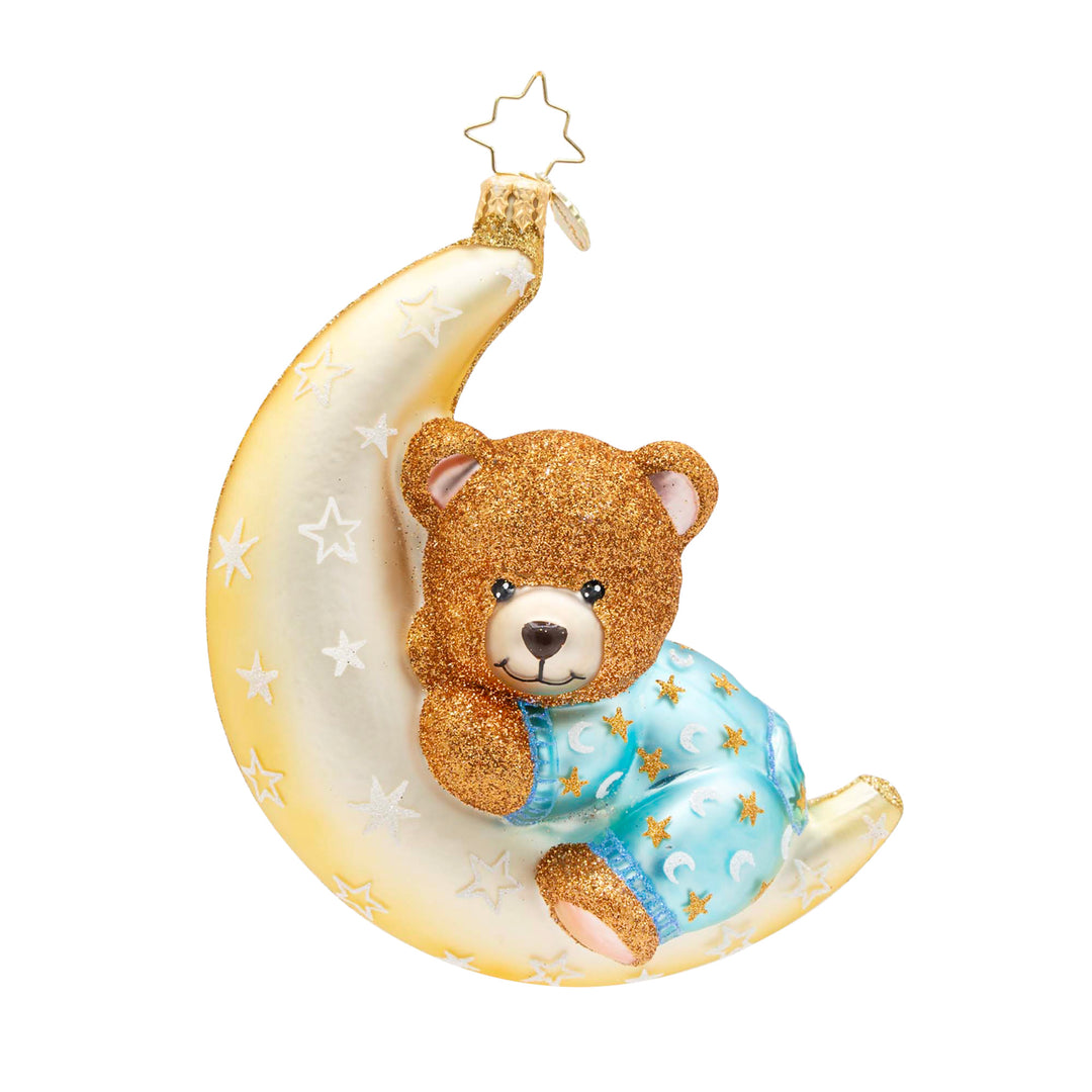 Blue Moonlight Snuggles. Ornament of a golden moon with white stars holding up a sparkly brown bear wearing blue pajamas. View of front