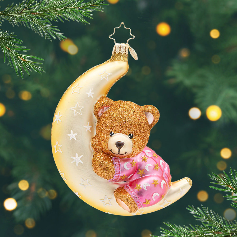 Pink Moonlight Snuggles. Ornament of a golden moon with white stars holding up a sparkly brown bear wearing pink pajamas. View of front