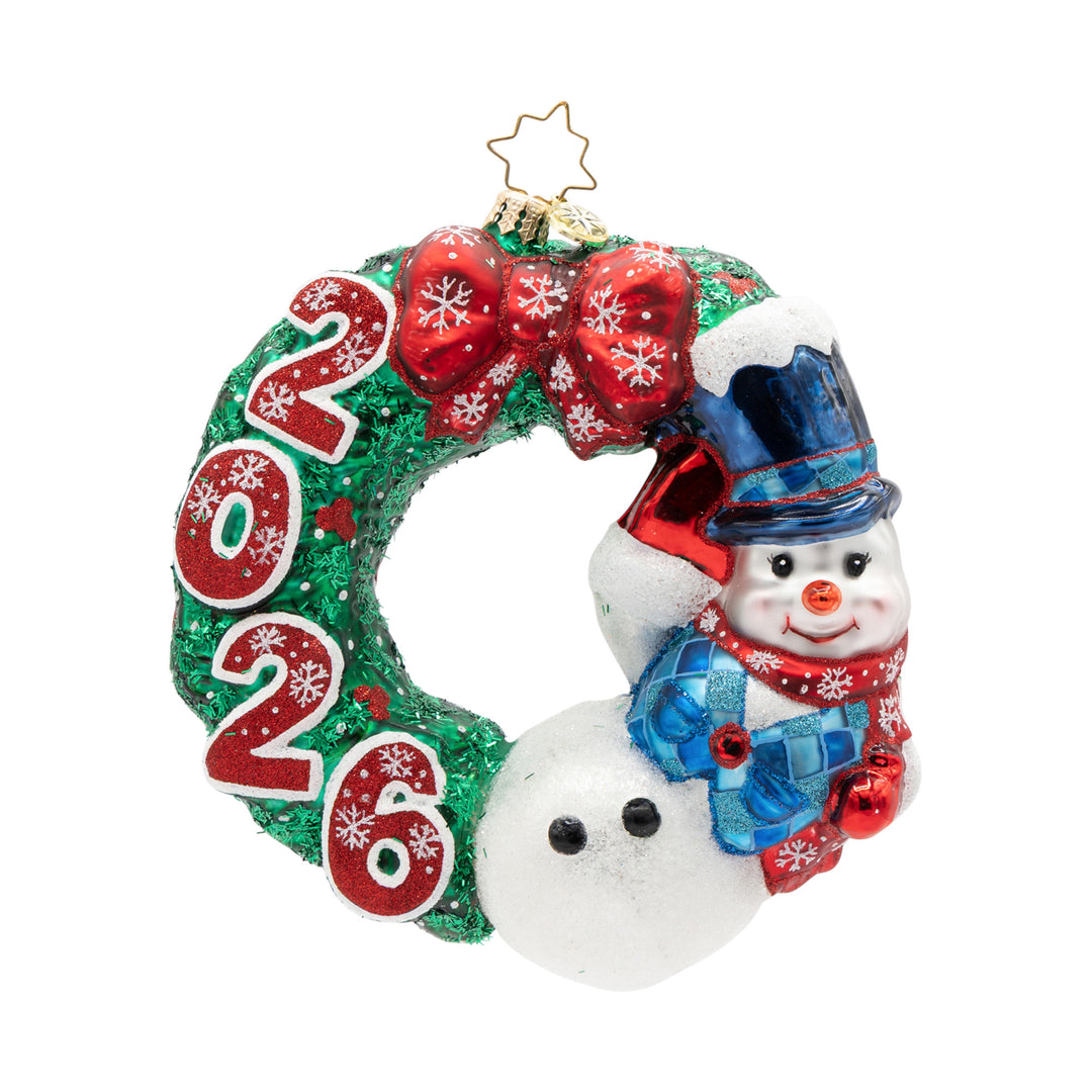 A Flurry of Cheer – 2026. A green wreath with 2026 in red on the left side and a snowman in a blue vest and top hat on the right and a red bow on top of the wreath. View of front