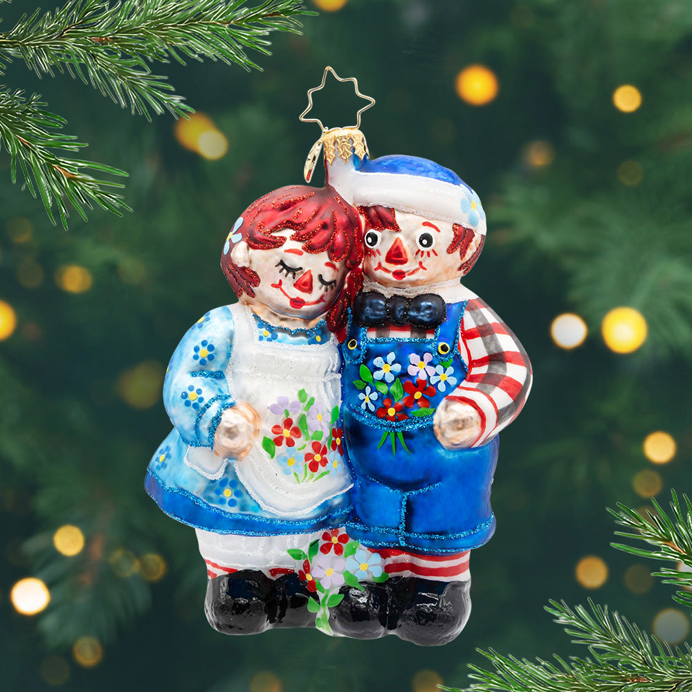 Raggedy Friends. Ornament of Raggedy Ann and Andy in blue outfits with flowers hugging each other. View of front