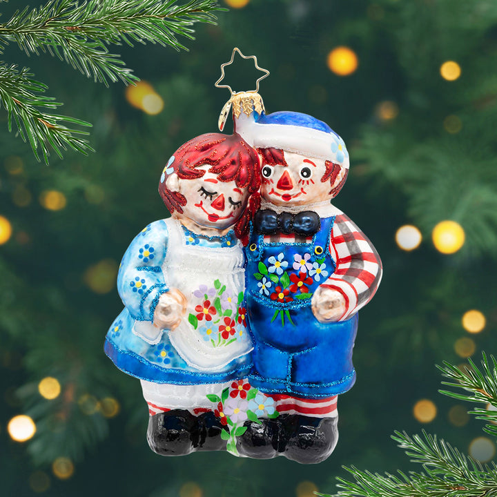 Raggedy Friends. Ornament of Raggedy Ann and Andy in blue outfits with flowers hugging each other. View of front