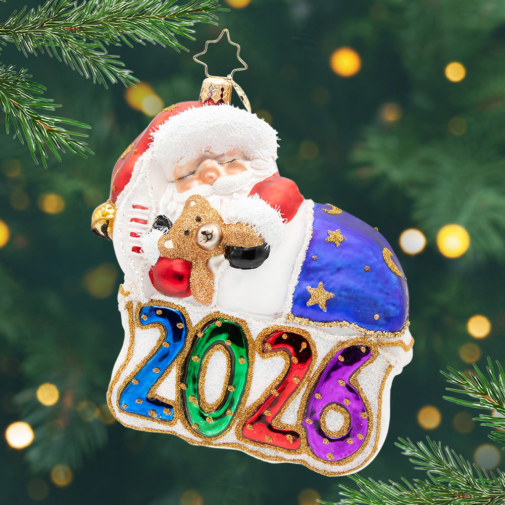 2026 Sweet Dreams. Ornament of Santa sleeping on top of the numbers 2026 while wrapped in a blue blanket and holding a teddy bear. View of front