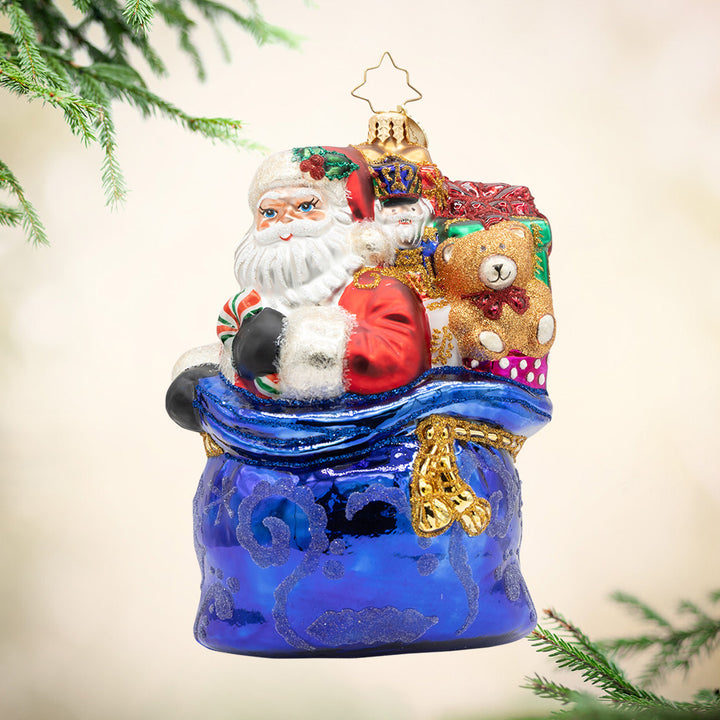 Kris Kringle’s Bundle. Ornament of Santa inside a giant blue bag with other presents. View of front.