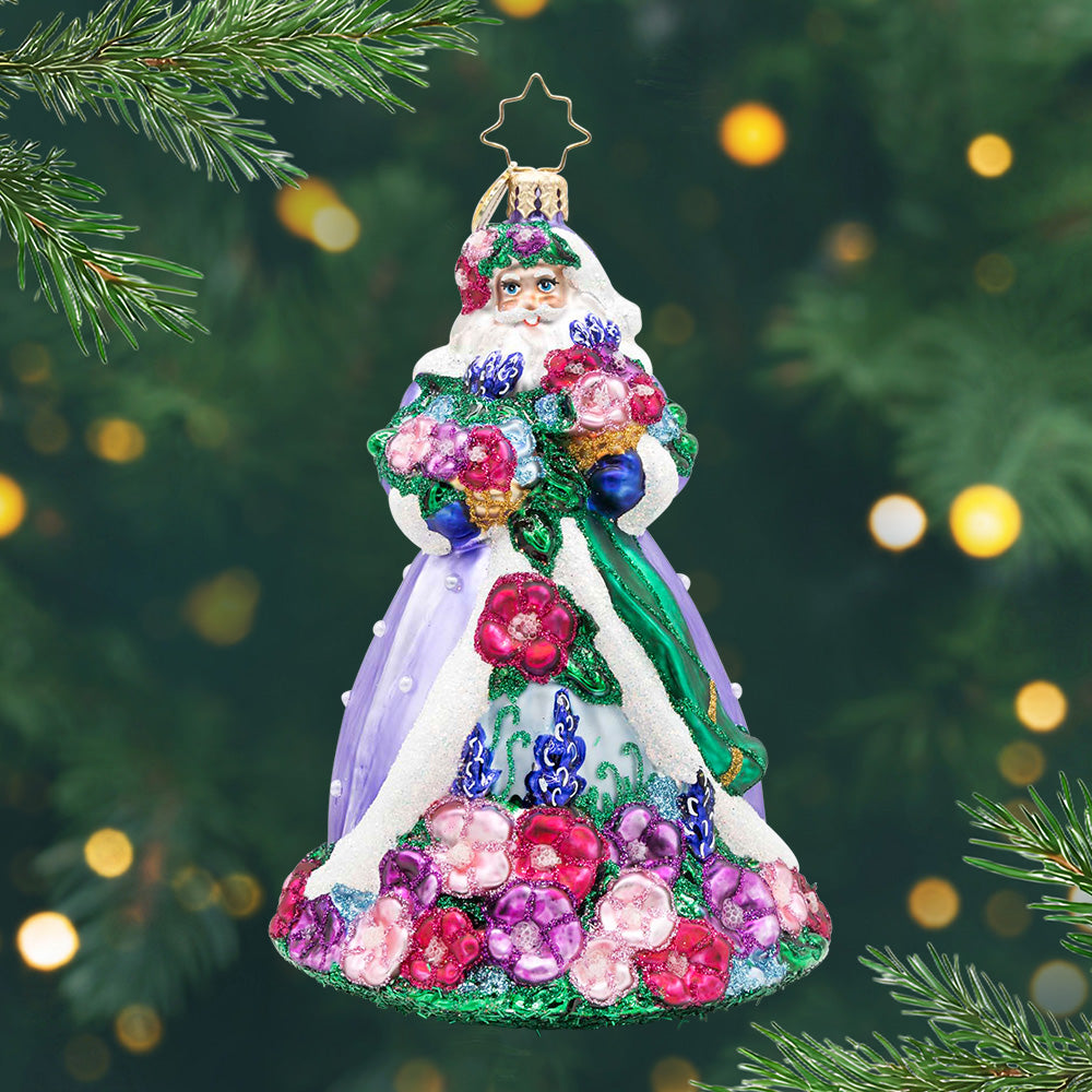 Santa in Full Bloom. Ornament of Santa dressed in a lavender and soft rose colored robe while carrying baskets overflowing with pansies, lavender, and hydrangeas with vine patterns and floral accents. View of front