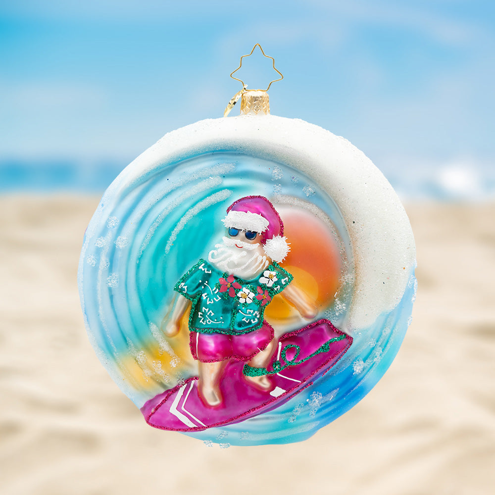 Surf’s Up, St. Nick! Ornament of Santa in a blue tropical shirt while wearing pink shorts and hat, riding on a pink surfboard inside a blue sparkling wave. View of front