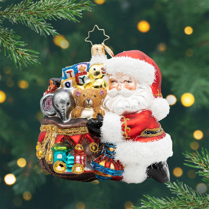 Santa’s Toyland Treasures. Santa ornament of him hugging a brown bag full of different toys include an elephant, teddy bear, blocks and a train. View of front.