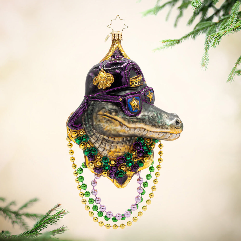 Gator Gras. Ornament. Alligator head wearing yellow, green, and purple beaded necklaces with a black and purple hat and glasses. View of front