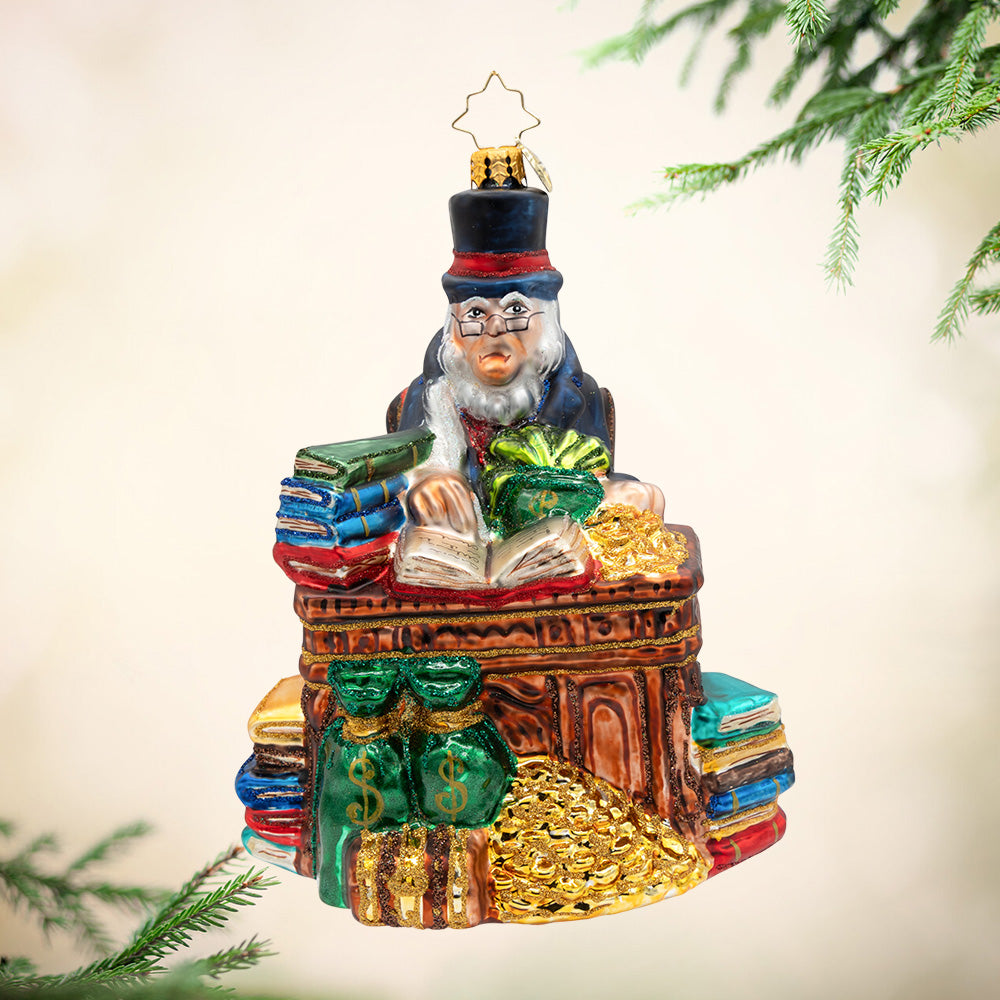 Tales from the Counting Desk. Ornament of Mr. Scrooge at his desk surrounded by ledgers, coins, and towering stacks of books. View of front