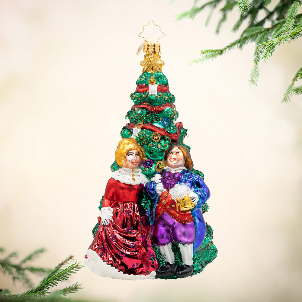 Festive Fezziwigs. An ornament of a decorated tree witih Mr. and Mrs. Fezziwig standing in front while dressed in bright colors of red, blue, and purple. View of front