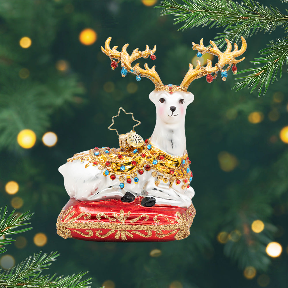 The Yuletide Jewel. Ornament of a white dear on top of an embroidered crimson pillow, adorned in raised golden jewelry and radiant gemstone hues. With glittering tassels and bejeweled antlers. View of front
