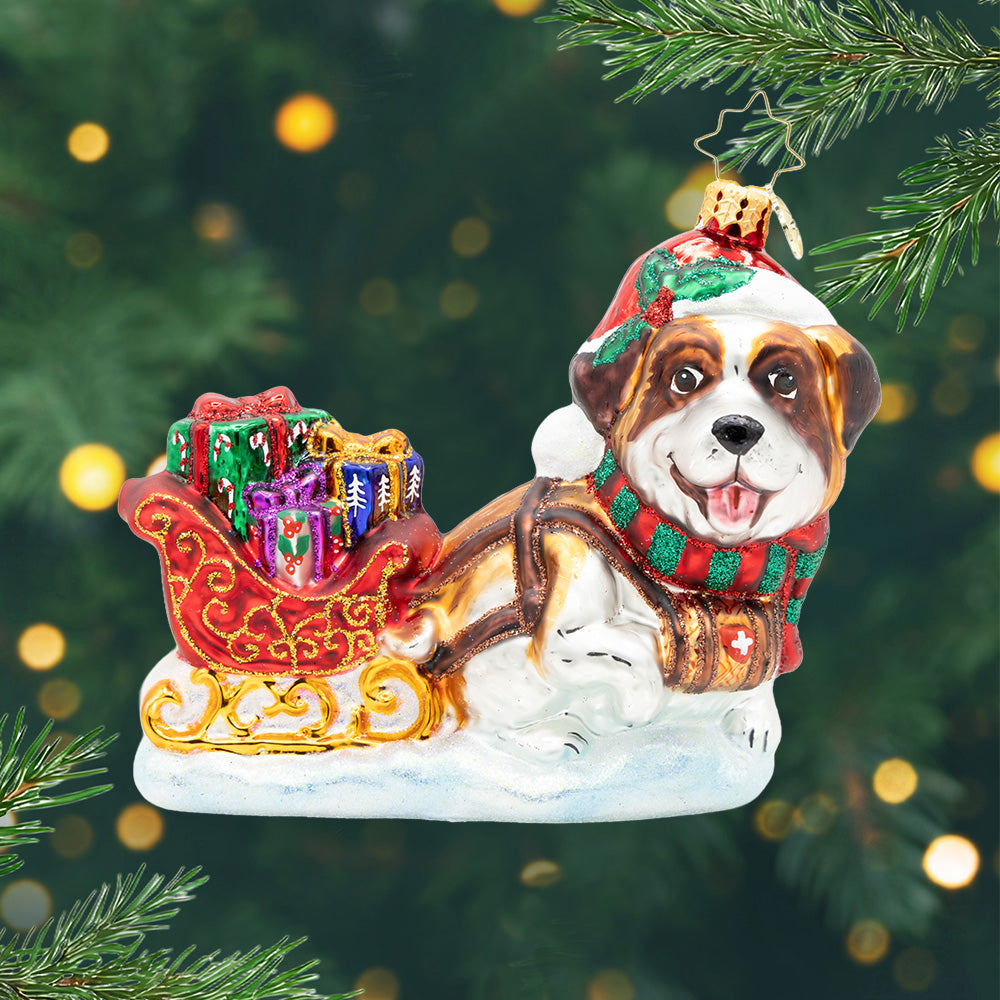North Pole Pawtrol. Ornament of a brown and white St. Bernard pulling a red and old detailed sleigh full of presents in colors of pink, green and blue. View of front.